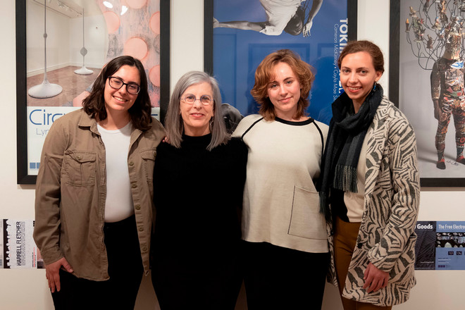 Four women, including Gallery Director Sharon McConnell, stand smiling in front of framed posters in the Poster Archives exhibit. The group includes three graphic design interns who contributed to the catalogue and exhibition design.