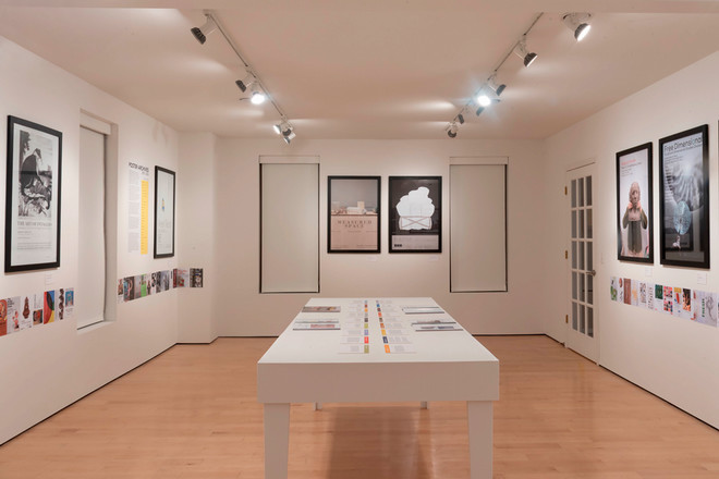 A quiet, wide-angle view of the Cohen Gallery featuring framed posters along the walls and a central white table displaying catalogue materials and timelines. Posters include minimalist designs and thematic artwork, with clean lighting and modern gallery styling.