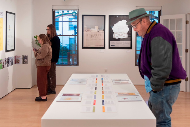 Three visitors examine the Poster Archives 2001–2024 exhibit at the Cohen Gallery. One man in a green fedora and purple jacket looks at the catalog display table in the foreground, while a couple in the background views wall posters and printed materials.