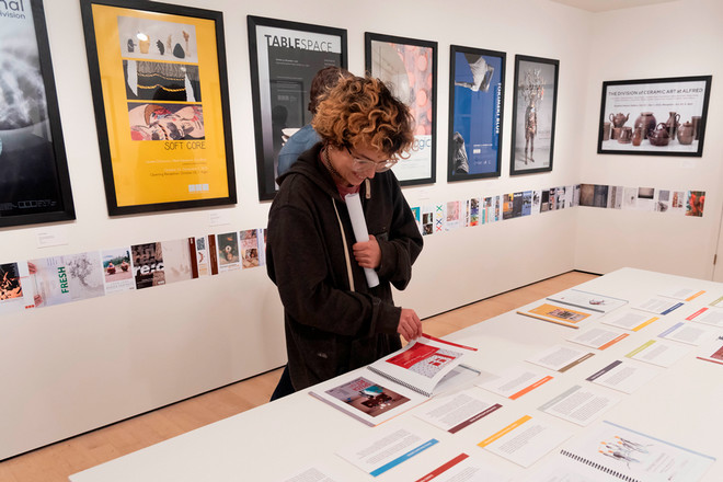 A visitor flips through the Poster Archives 2001–2024 catalogue on display at a central white table. Behind them, framed posters such as “Soft Core,” “Table Space,” and “Nick Cave” line the walls, with a timeline of smaller exhibition cards running along the wall base.