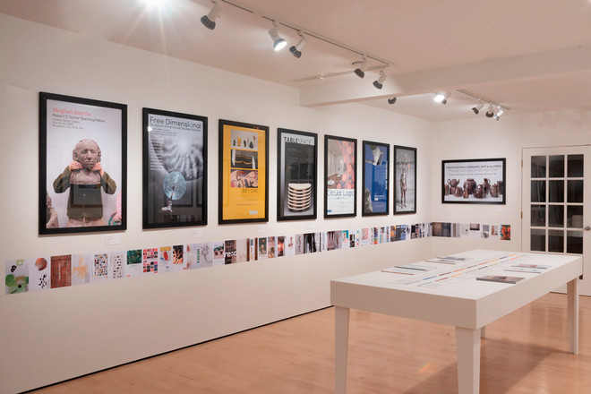 A gallery wall at the Poster Archives 2001–2024 exhibit featuring framed posters including “Meghan Smythe,” “Free Dimensional,” “Soft Core,” “Table Space,” and “Nick Cave.” A white display table with catalogs and text panels runs parallel to the poster wall, and a timeline of small prints lines the lower wall edge.
