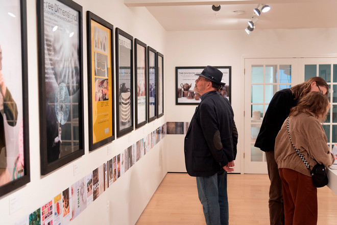 Three visitors examine a wall of framed exhibition posters at the Cohen Gallery. A man in a dark jacket and fedora closely inspects a poster titled “Soft Core,” while two others lean over a table to look at the catalog and print materials on display.