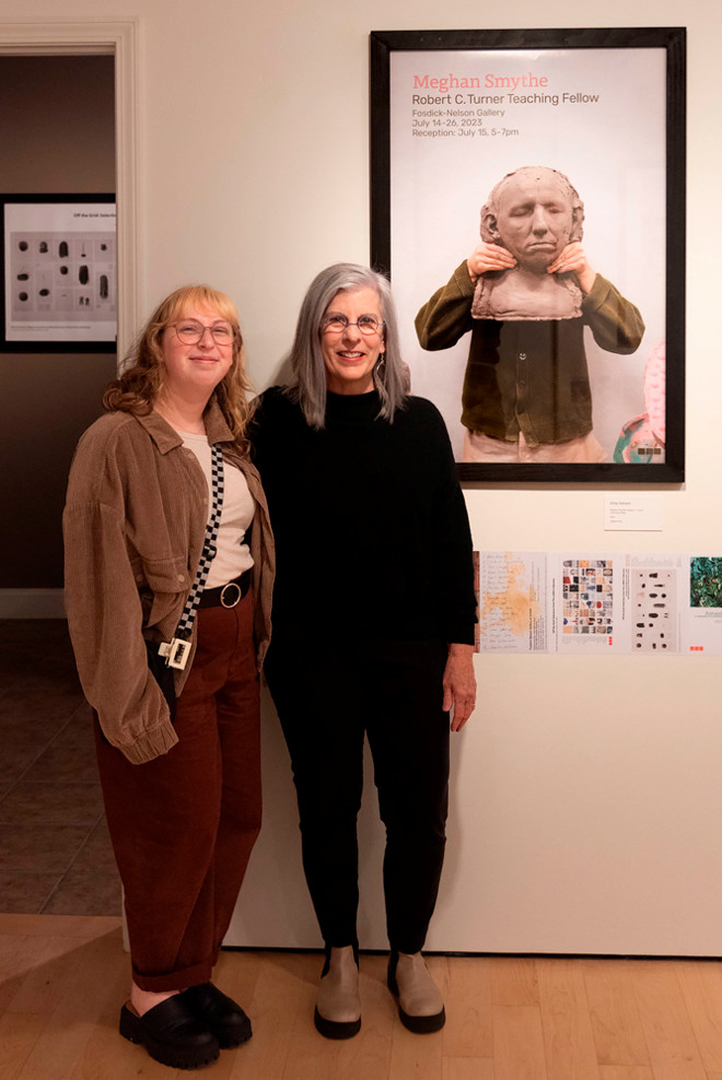 Gallery Director Sharon McConnell stands with intern Emily Sierzant in front of a framed exhibition poster for “Meghan Smythe: Robert C. Turner Teaching Fellow.” They smile for the photo, positioned near a wall lined with printed poster thumbnails and project descriptions.