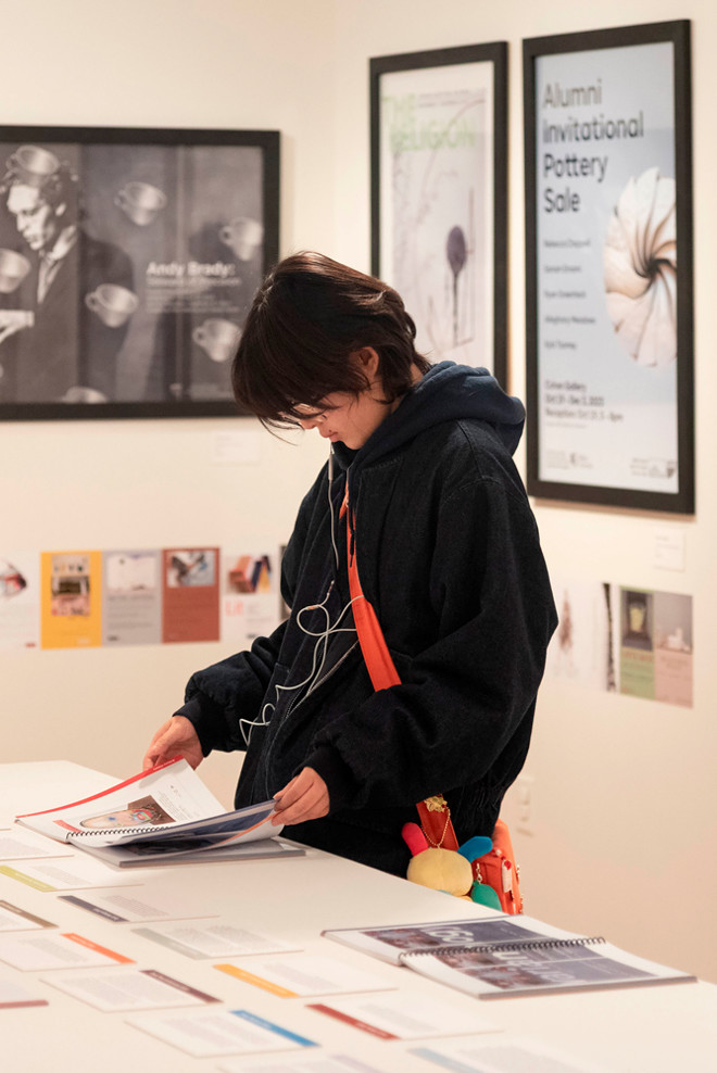 A visitor in a dark hoodie with headphones browses through the Poster Archives 2001–2024 catalogue on a display table. Behind them, framed posters including “Andy Brady,” “The Religion,” and “Alumni Invitational Pottery Sale” are mounted on the gallery wall.
