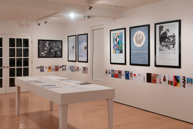 A side view of the Cohen Gallery showing framed posters including “Errant Landscapes,” “Alfred/Düsseldorf MFA in Painting,” and “The Art of Intaglio.” A central table displays open catalogues and timeline documents, while small posters line the base of the walls throughout the exhibit.