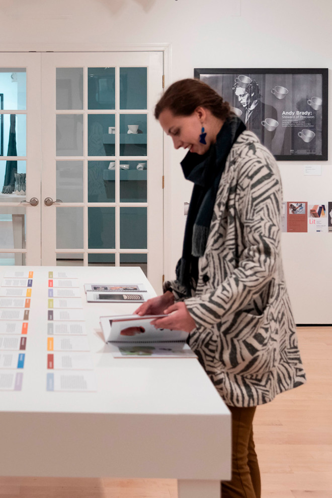 Natalie Burns, a former gallery intern, flips through the Poster Archives catalogue at a display table in the Cohen Gallery. In the background, a framed poster for “Andy Brady: Steward of Ironsicon” is mounted on the wall near a set of glass doors.