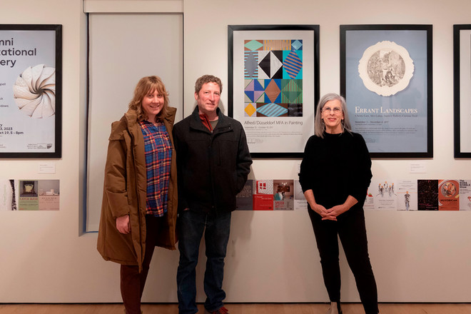 Three people pose in front of a gallery wall displaying posters for “Alfred/Düsseldorf MFA in Painting,” “Errant Landscapes,” and the “Alumni Invitational Pottery Sale.” Gallery Director Sharon McConnell stands on the right, smiling beside the guests.