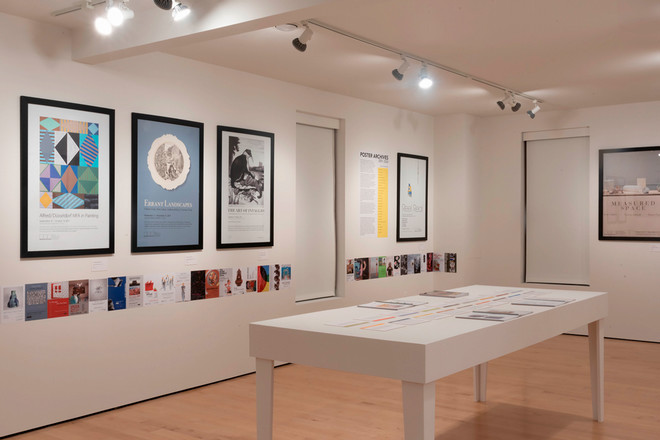A wide shot of the Cohen Gallery showing framed posters including “Alfred/Düsseldorf MFA in Painting,” “Errant Landscapes,” and “The Art of Intaglio.” Below the posters is a timeline of smaller printed cards. A white table in the center displays open exhibition catalogues and color-coded layout strips.
