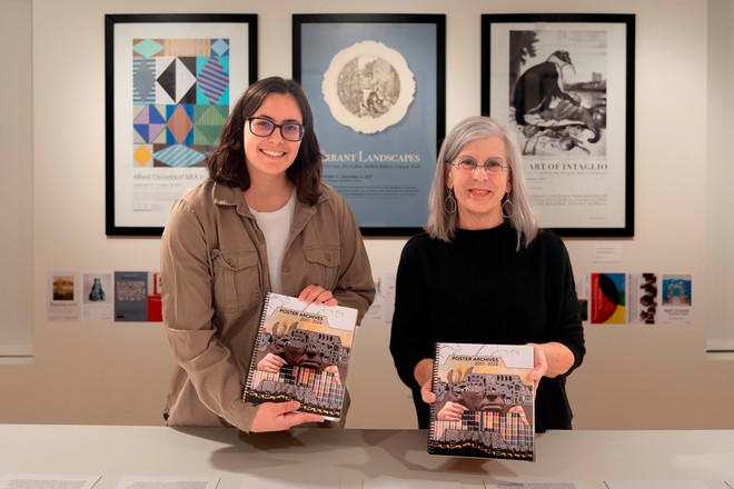 Gallery Director Sharon McConnell and graphic designer Jemma Lindquist stand side-by-side holding copies of the Poster Archives 2001–2024 catalogue. Behind them are framed posters including “Alfred/Düsseldorf MFA in Painting,” “Errant Landscapes,” and “The Art of Intaglio.”
