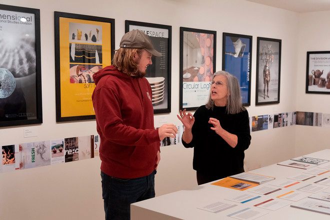 Gallery Director Sharon McConnell speaks with a visitor wearing a red hoodie and cap in front of a display of framed posters, including “Soft Core,” “Table Space,” “Circular Logic,” and “Nick Cave.” The exhibition catalogue and timeline materials are visible on the table beside them.