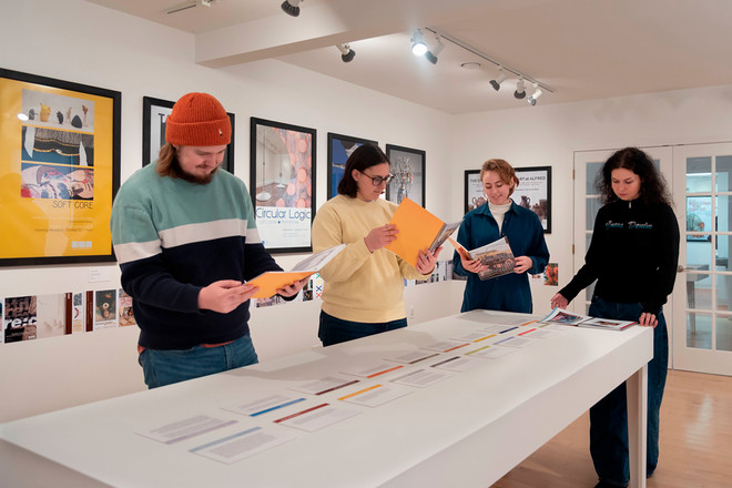 Four gallery interns review catalog layouts and poster materials on a long display table at the Cohen Gallery. Behind them, framed posters such as “Soft Core,” “Circular Logic,” and “Nick Cave” hang on the wall. The interns hold documents and folders as they collaborate on the installation.