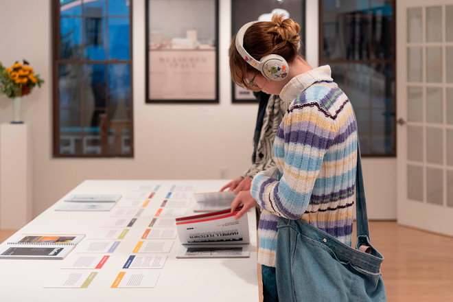 A visitor wearing colorful striped headphones and a pastel sweater browses through the Poster Archives 2001–2024 catalogue during the opening reception. Behind them, framed posters including “Measured Space” are displayed on the gallery wall, and layout sheets are spread across the central display table.