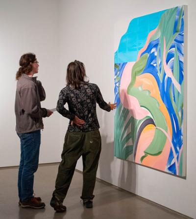 Two people stand in a gallery, closely observing a large colorful abstract painting composed of flowing shapes and bold brushstrokes.