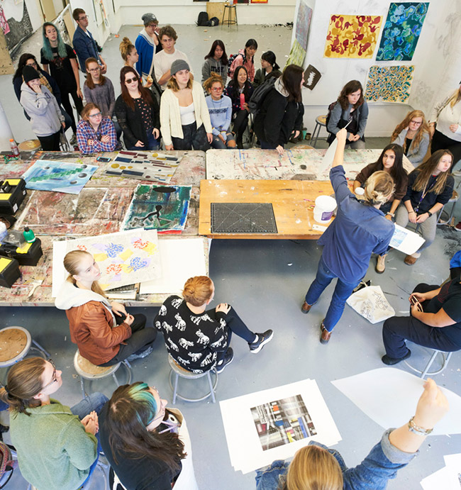A large group of art students gathers in a bright studio filled with tables covered in paint and materials. A faculty member at the front raises an arm while leading a critique or demonstration, as students sit and stand around, looking at artwork laid out on the tables and floor.