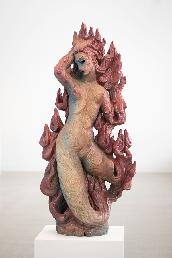 A sculpted ceramic figure of a nude woman emerging from swirling flame-like shapes, displayed on a white pedestal in a gallery space.