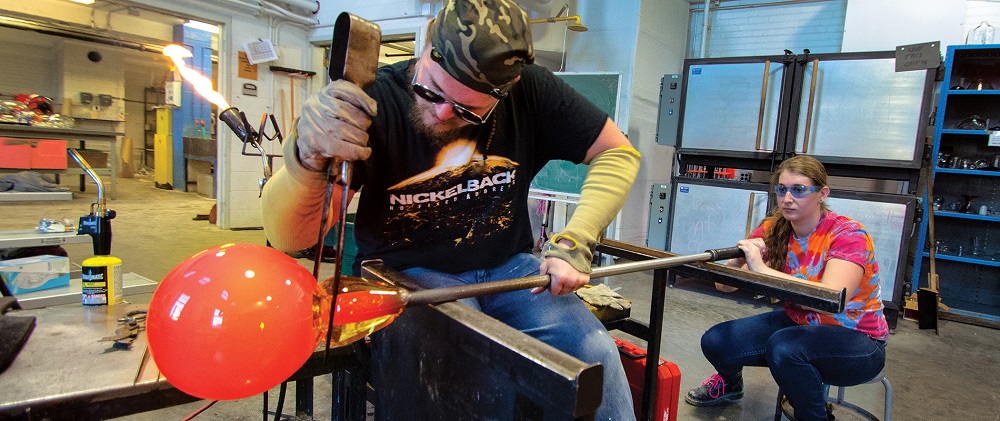 student clamping glass off of a pipe while another student blows to inflate the piece