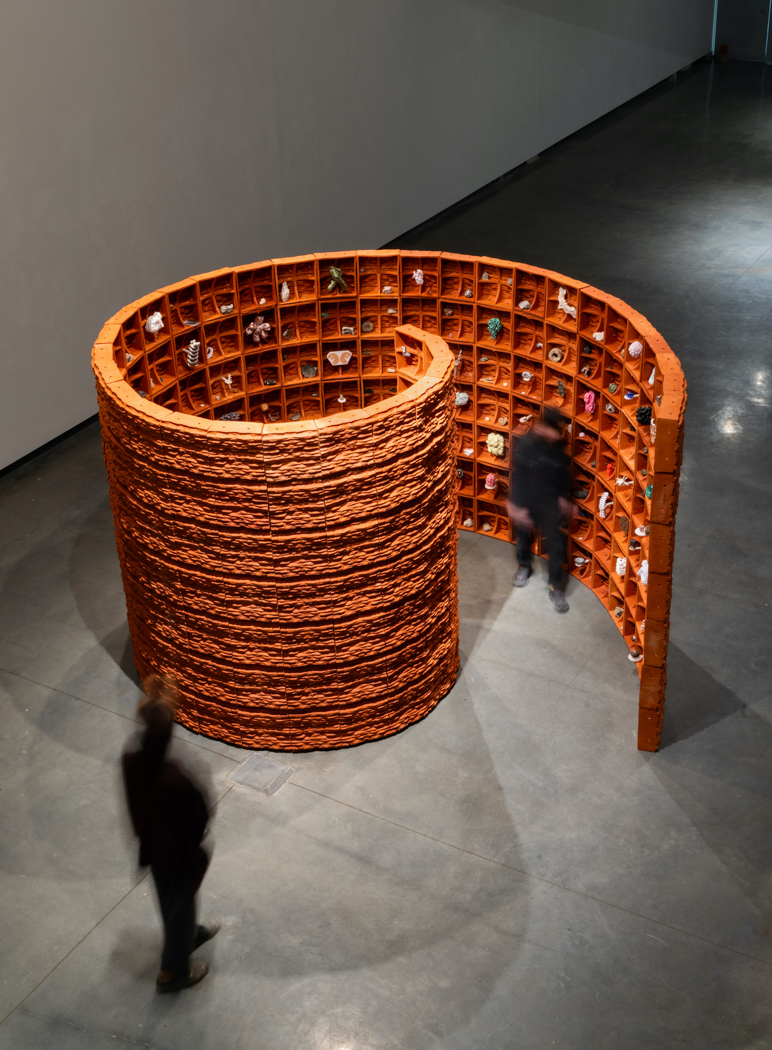 A large spiral-shaped art installation made of reddish-brown, clay-like material stands in a modern gallery space. The cylindrical structure coils inward, forming a walkable path lined with small cubbyholes embedded in its textured walls. Each cubby contains colorful miniature objects or figurines, creating a vibrant contrast against the earthy tones. Two visitors stroll nearby, highlighting the scale and inviting nature of the immersive sculpture.
