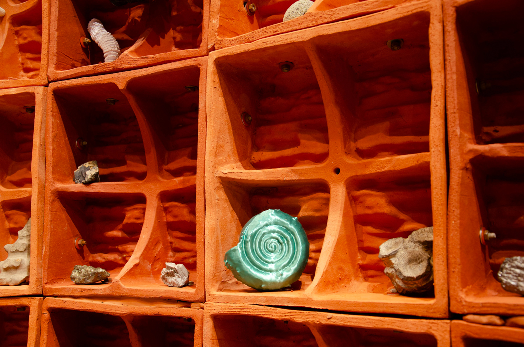 Close-up of an orange clay display case with compartments holding rocks, fossils, and a green spiral-shaped object.