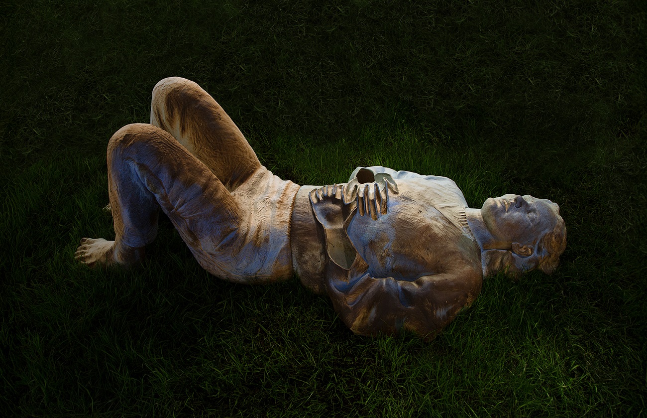 Wooden sculpture of a person lying on grass with bent legs and hands on chest, illuminated to show carved details.