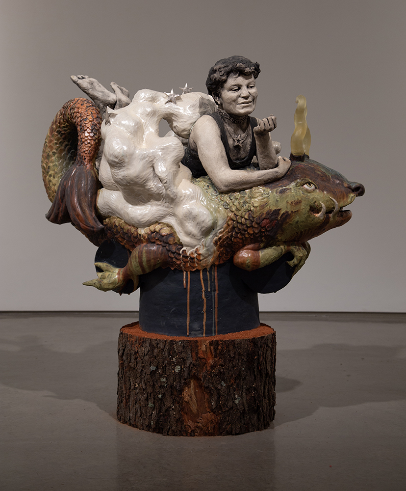 Sculpture of a person resting on a colorful fish-like creature, surrounded by white cloud shapes and mounted on a painted tree trunk base.