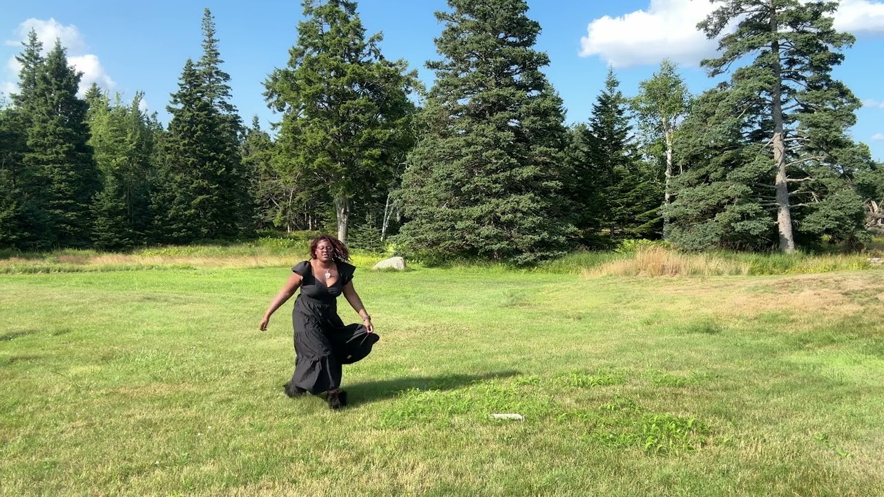 Person in a black dress walking across a grassy field bordered by tall evergreen trees under a partly cloudy blue sky.