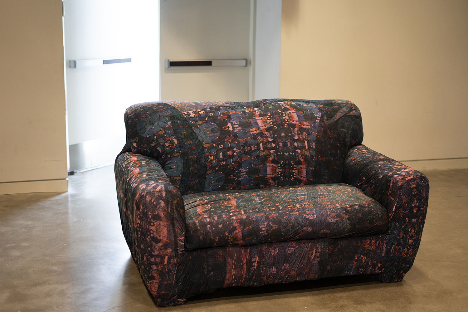 Patterned loveseat with abstract blue, red, and orange design in a room with concrete floor and white walls; a door with a push bar is visible in the background.