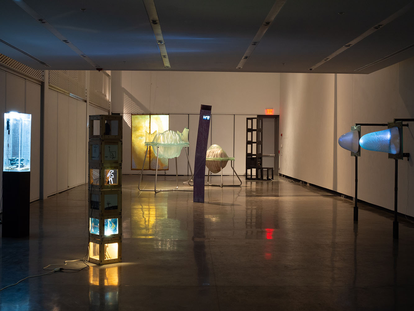 Dimly lit gallery with illuminated sculptures, including a tall column with screens, a translucent shell-like piece, and conical wall lights.