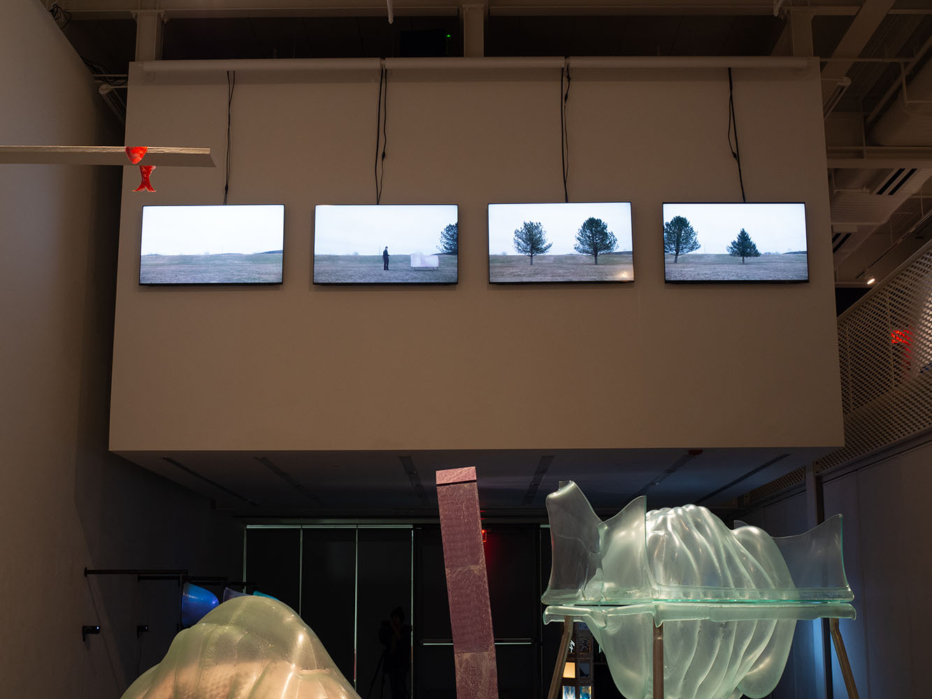 Gallery installation with four screens showing outdoor scenes and abstract translucent sculptures in the foreground.