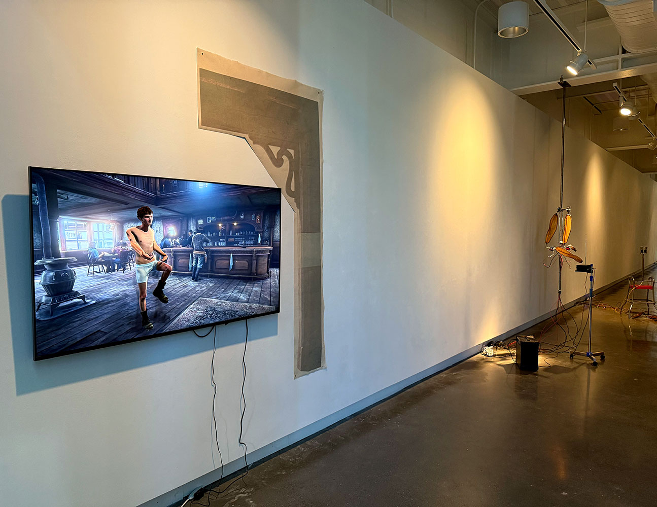 Gallery wall with a screen showing a tavern scene from a digital artwork, exposed drywall, and a nearby light-based installation with objects on stands.