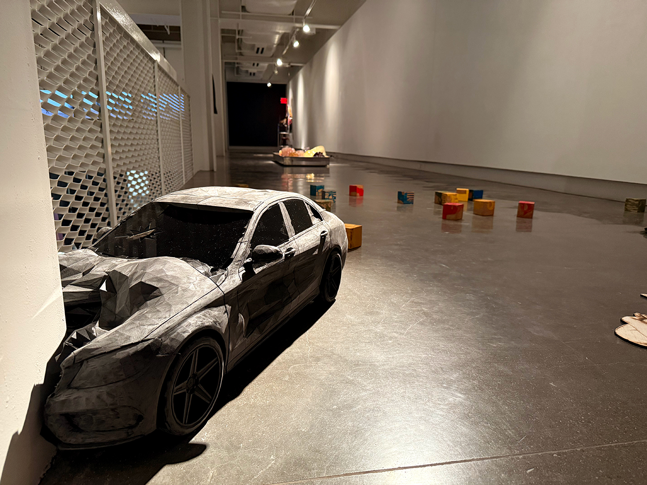 Crashed black car model against a white gallery wall, with scattered colorful blocks and two human-like sculptures on the floor.