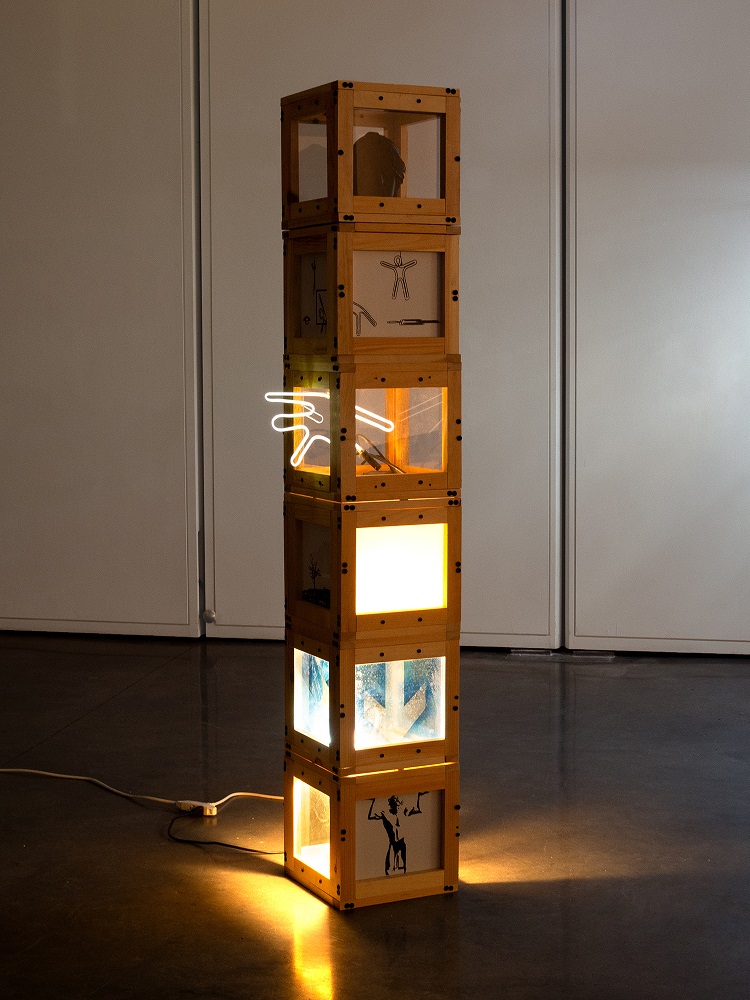 Tall wooden sculpture of stacked illuminated cubes with abstract designs, glowing warmly in a gallery space