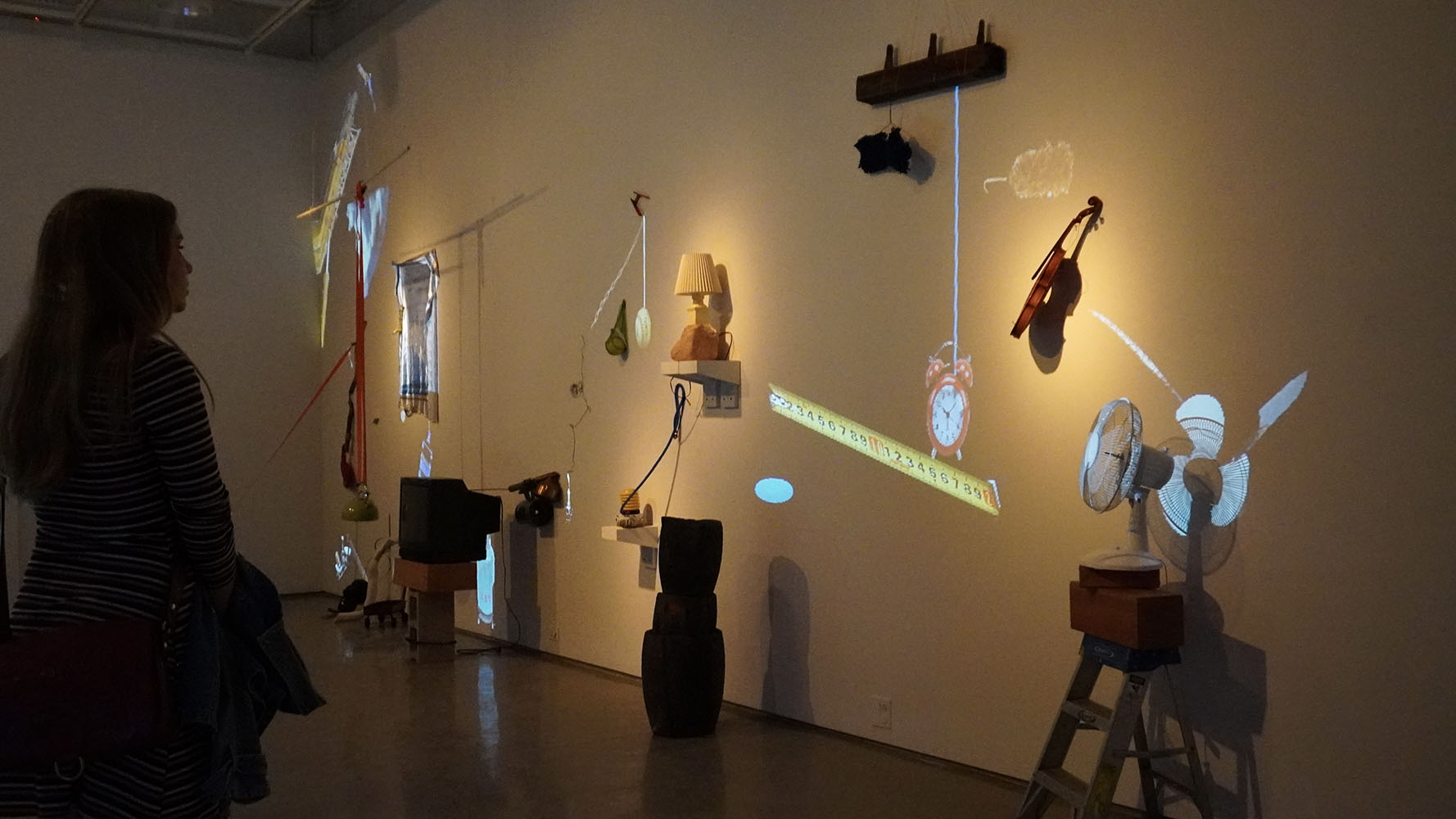 Person viewing a wall-mounted art installation with illuminated everyday objects like a violin, fan, and lamp in a gallery setting.