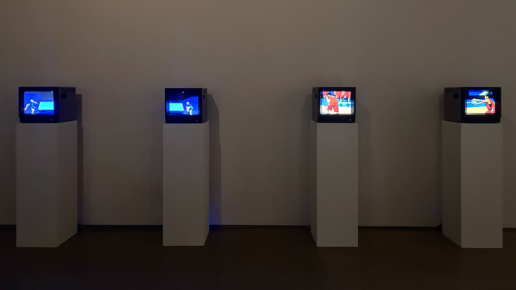 Four CRT TVs on white pedestals display sports scenes in a dimly lit gallery space.