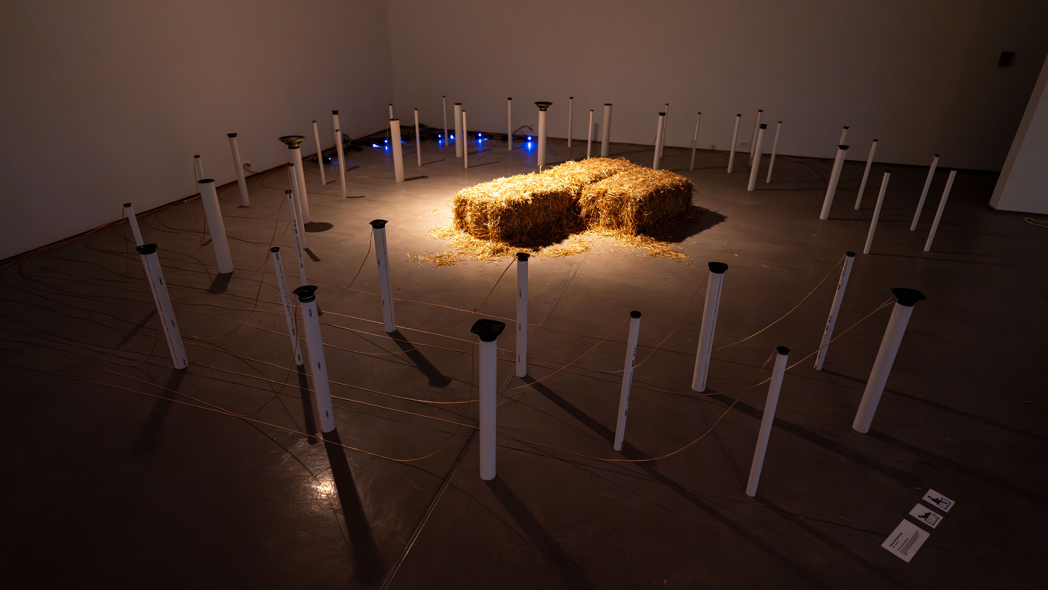 Hay bale installation with white poles and ropes, lit by blue lights.