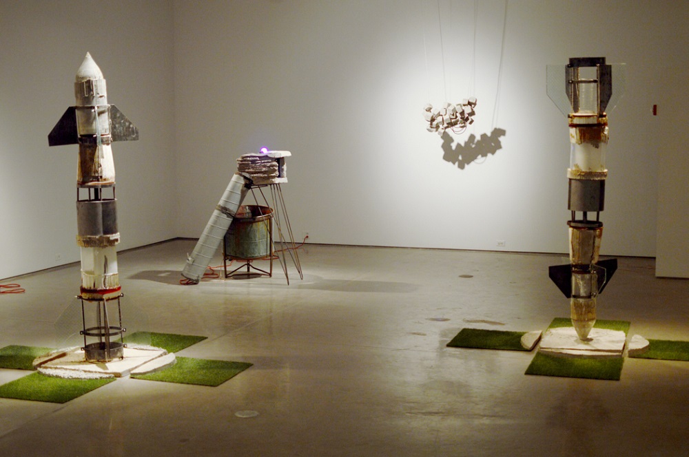 Three rocket-like sculptures on artificial grass in a gallery.