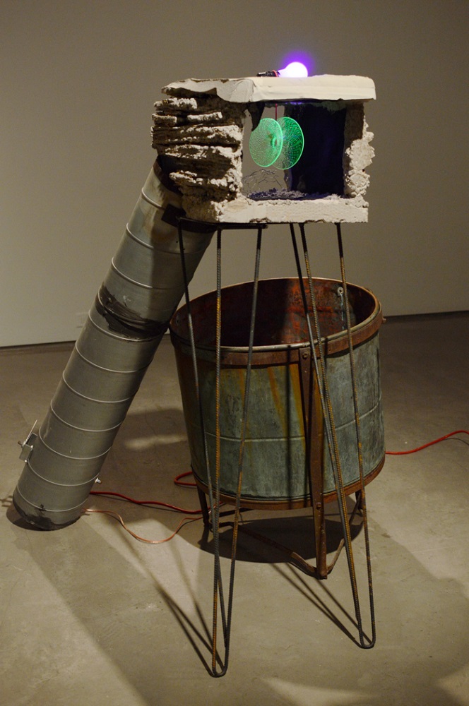Sculpture with a metal drum and concrete structure lit by green and purple lights.