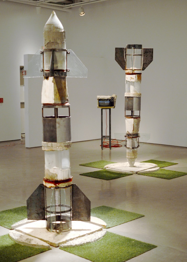 Two tall mixed media rocket sculptures on display in a gallery.