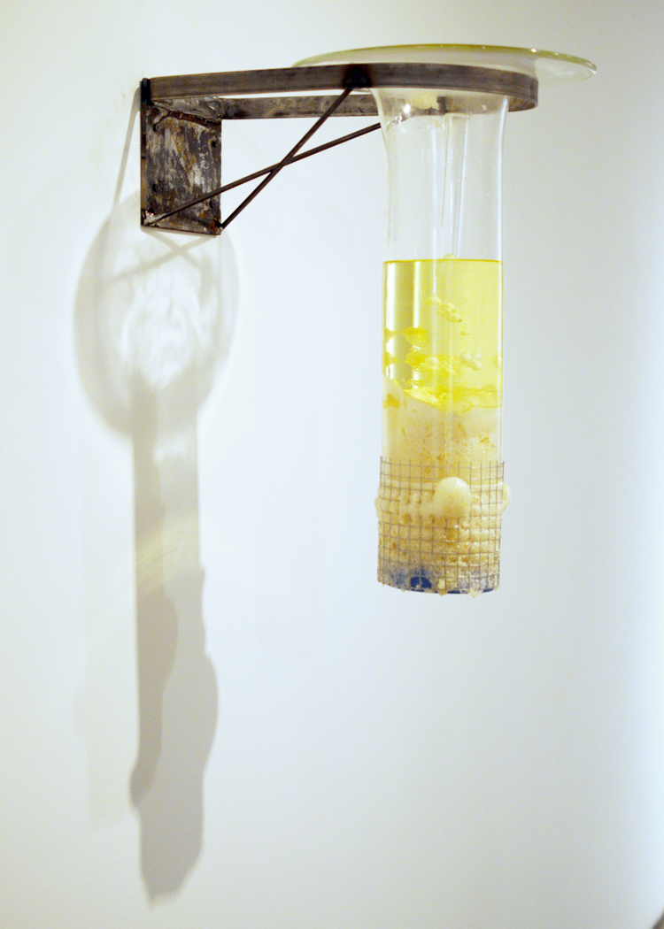 Wall-mounted glass cylinder with yellow liquid and objects at the bottom.