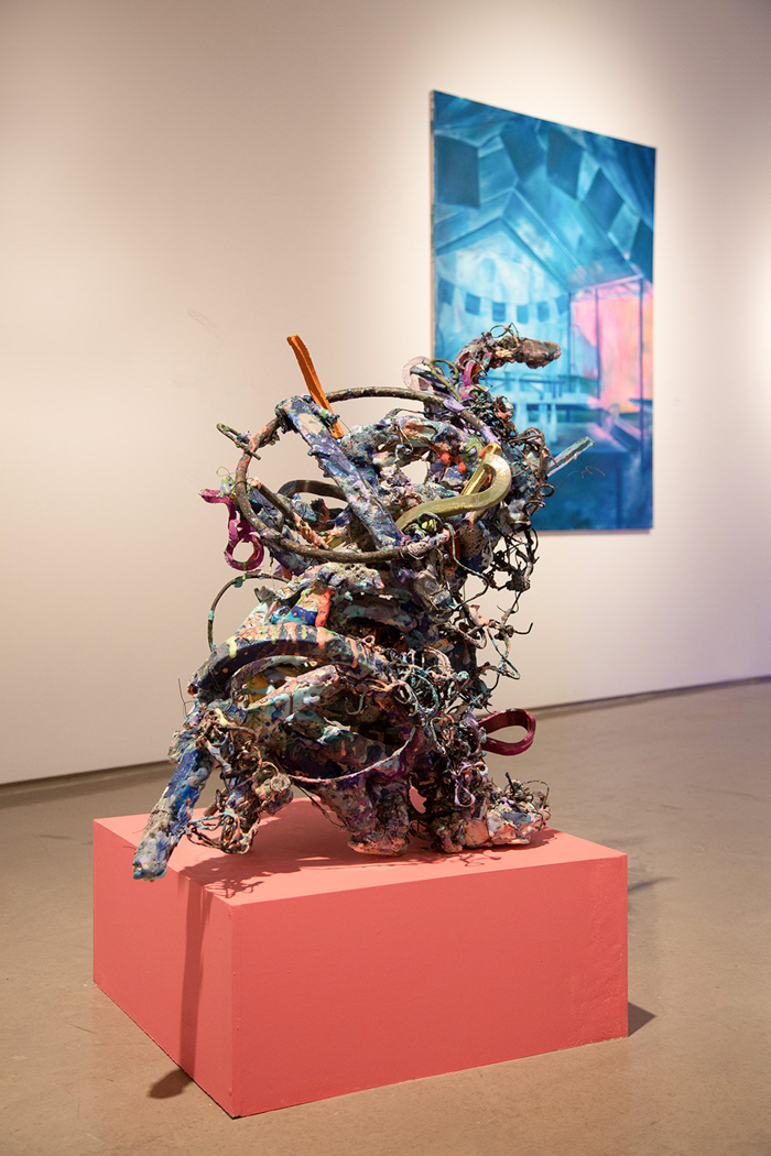 Multicolored abstract sculpture on a pink pedestal with a blue painting behind.