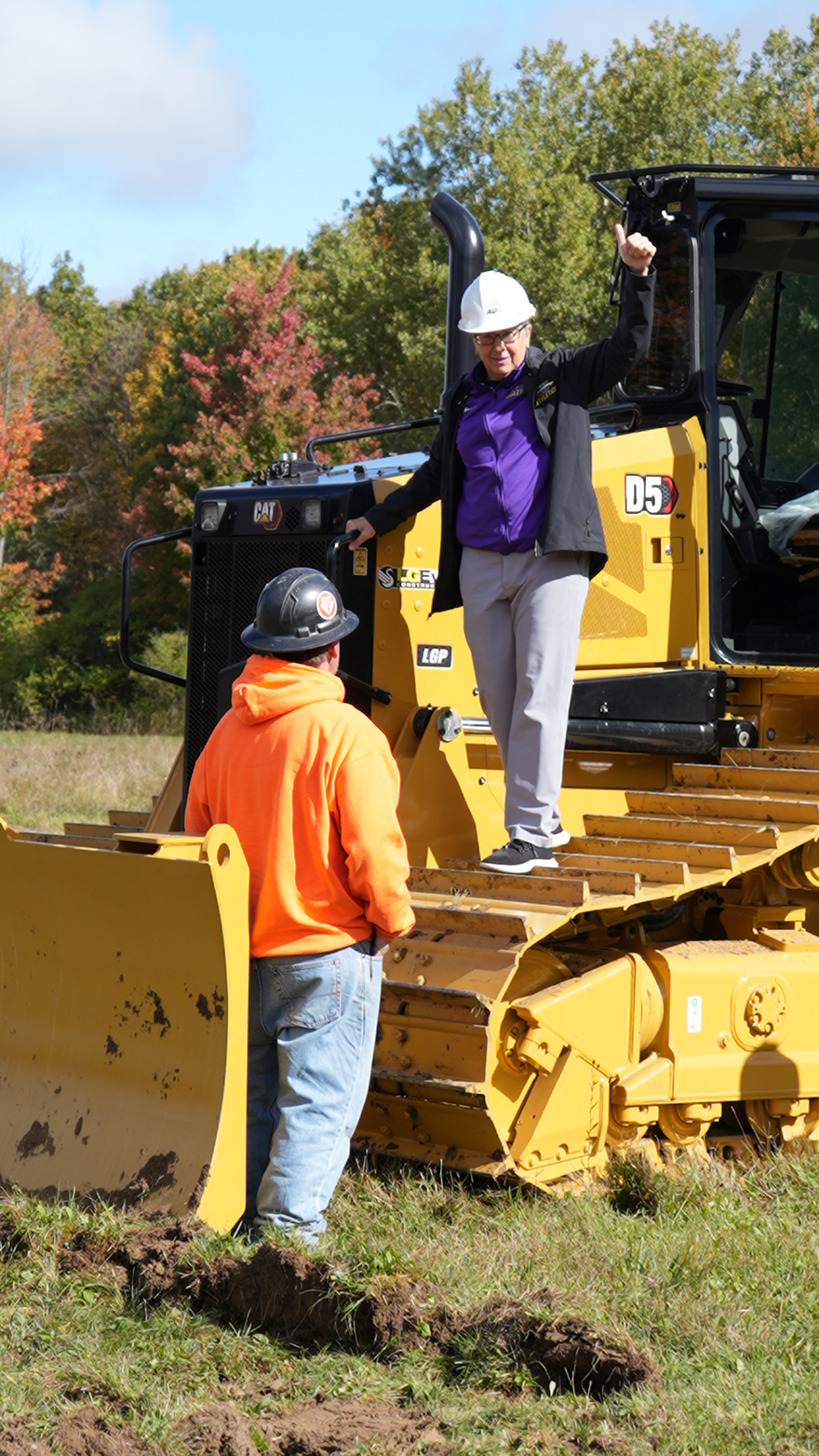 Person in a hard hat standing on a bulldozer blade giving a thumbs-up to another person