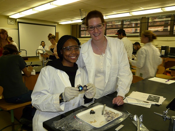 A student and instructor wearing lab coats and safety goggles smile while working on a dissection project in a classroom laboratory.