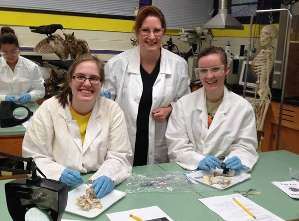 Two students and their instructor smile while working on a dissection activity in a psychology laboratory, wearing lab coats, gloves, and safety goggles.