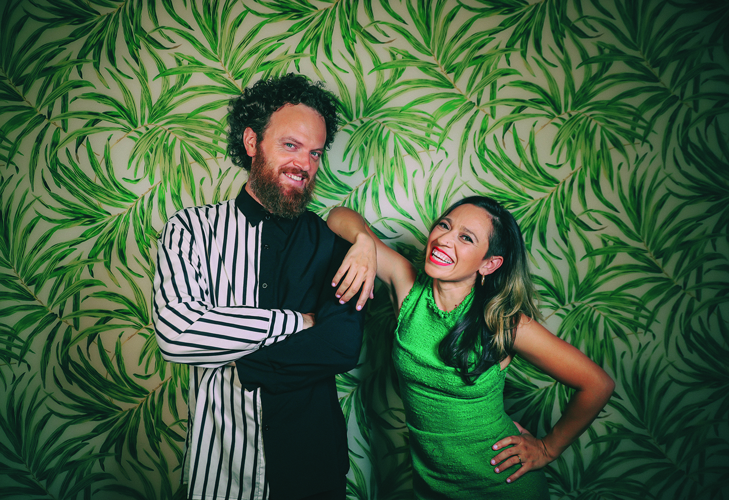 A large group of formally dressed musicians and guests pose together indoors after a performance, many holding drinks and smiling at the camera. They wear black-tie and concert attire in a bright lobby with columns, tiled floors, and soft overhead lighting.